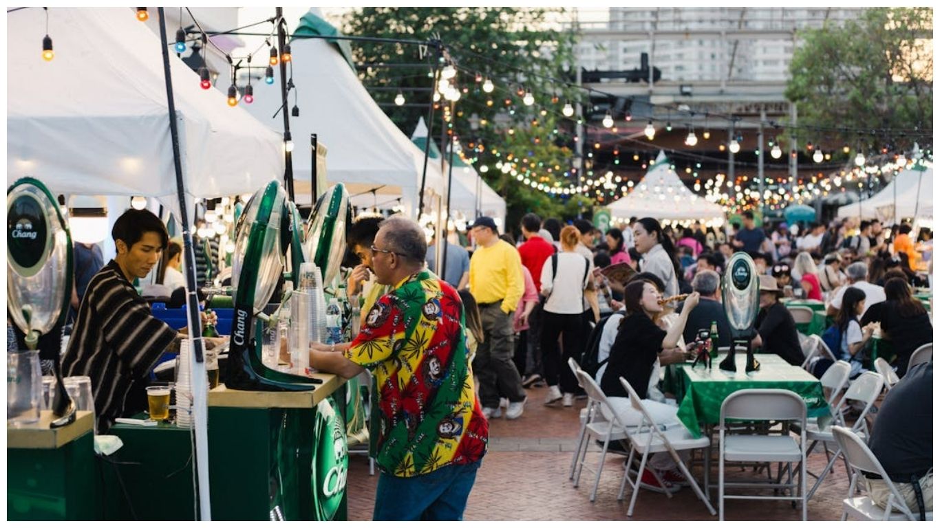 People dining at an outdoor festival in Thailand with Chang beer stalls and decorative string lights.