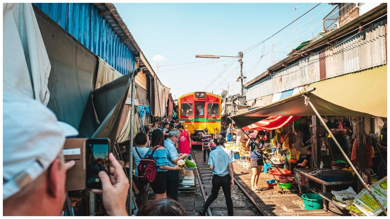 A colorful train passing through the crowded Maeklong Railway Market in Bangkok with tourists taking photos and vendors lining the tracks