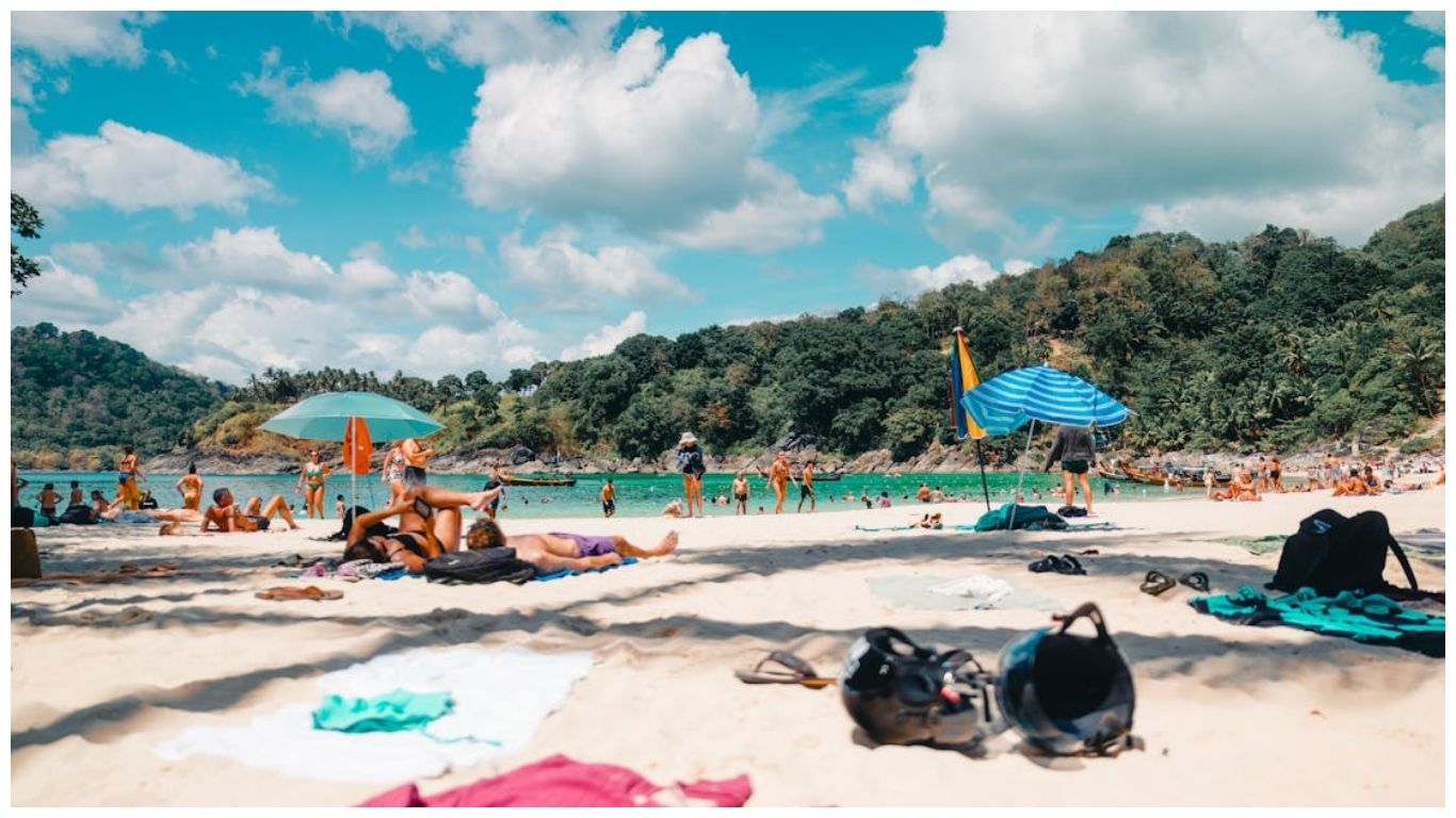 A sunny day at a crowded Phuket beach with tourists sunbathing, colorful umbrellas on white sand, and turquoise water against a backdrop of lush green hills