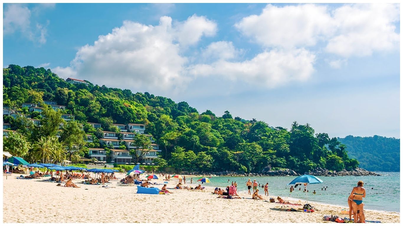 A beautiful landscape of a Phuket beach featuring white sand, clear turquoise water, and luxury resorts nestled on a lush green hillside under a blue sky