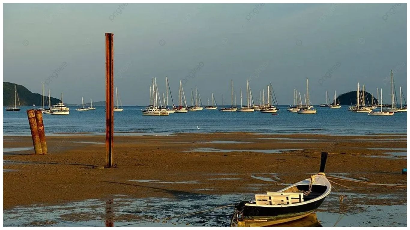 A peaceful beach in Phuket at low tide with a traditional Thai longtail boat in the foreground and several white sailboats anchored in the calm blue bay under a clear sky