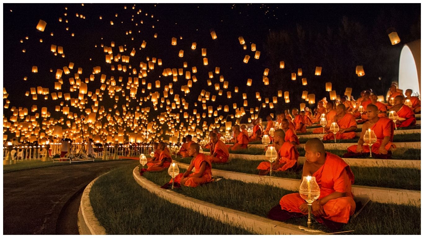 Thousands of sky lanterns floating in the dark night sky over Buddhist monks sitting in prayer during the Yi Peng Festival, a highlight of Thailand in November