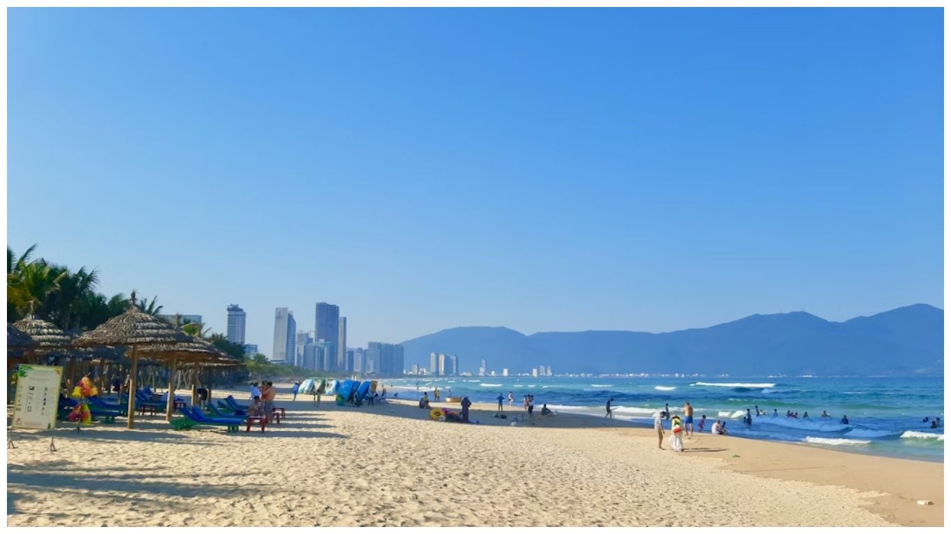 A sunny day at My Khe Beach in Da Nang featuring blue skies, thatched umbrellas, and travelers enjoying the sand and sea with the city skyline in the background, summarizing how many days in Da Nang to visit.