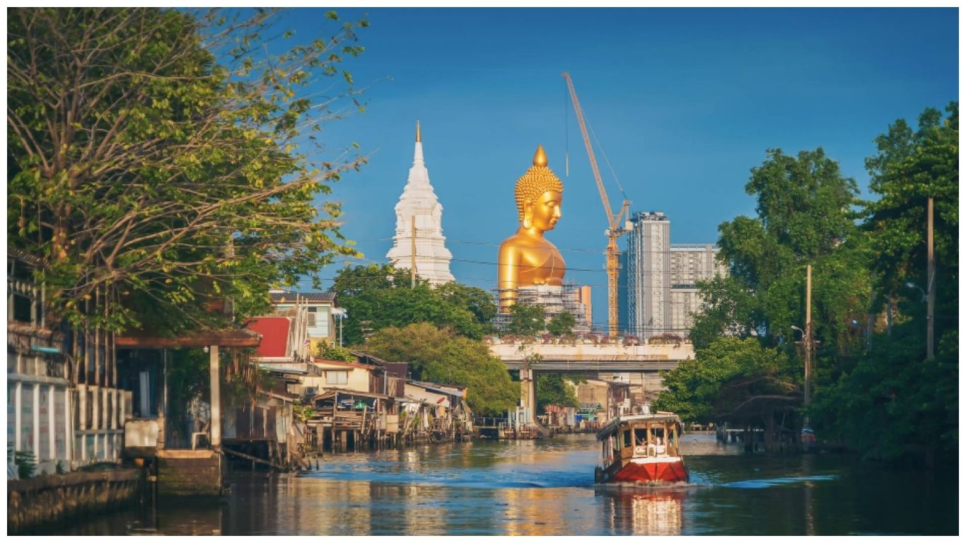 A scenic view of the Great Buddha of Wat Paknam Phasi Charoen rising above a Bangkok canal with a traditional longtail boat, representing the clear skies of Thailand in December weather.