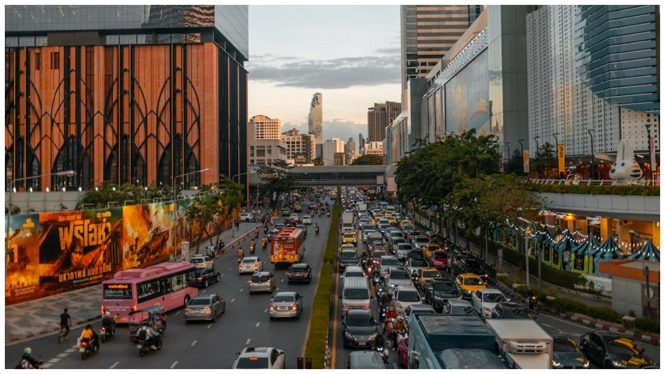 A high-angle view of a busy Bangkok street filled with cars, pink buses, and motorcycles against a backdrop of modern skyscrapers and the city skyline at dusk