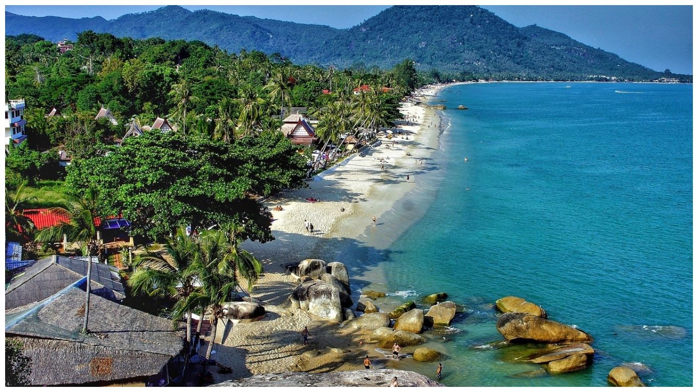 Panoramic view of a white sand beach with turquoise water, large granite boulders, and lush green mountains in the background, Thailand