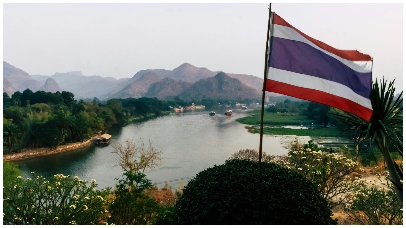 The Thai flag waving over the beautiful River Kwai with mountains in the background, a must-see for anyone exploring fun facts about Thailand