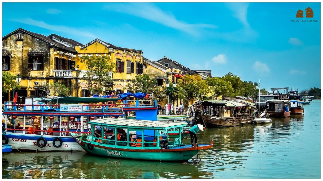 Bright yellow heritage buildings and colorful boats along the Thu Bon River in Hoi An Ancient Town, Vietnam, under a clear blue sky.