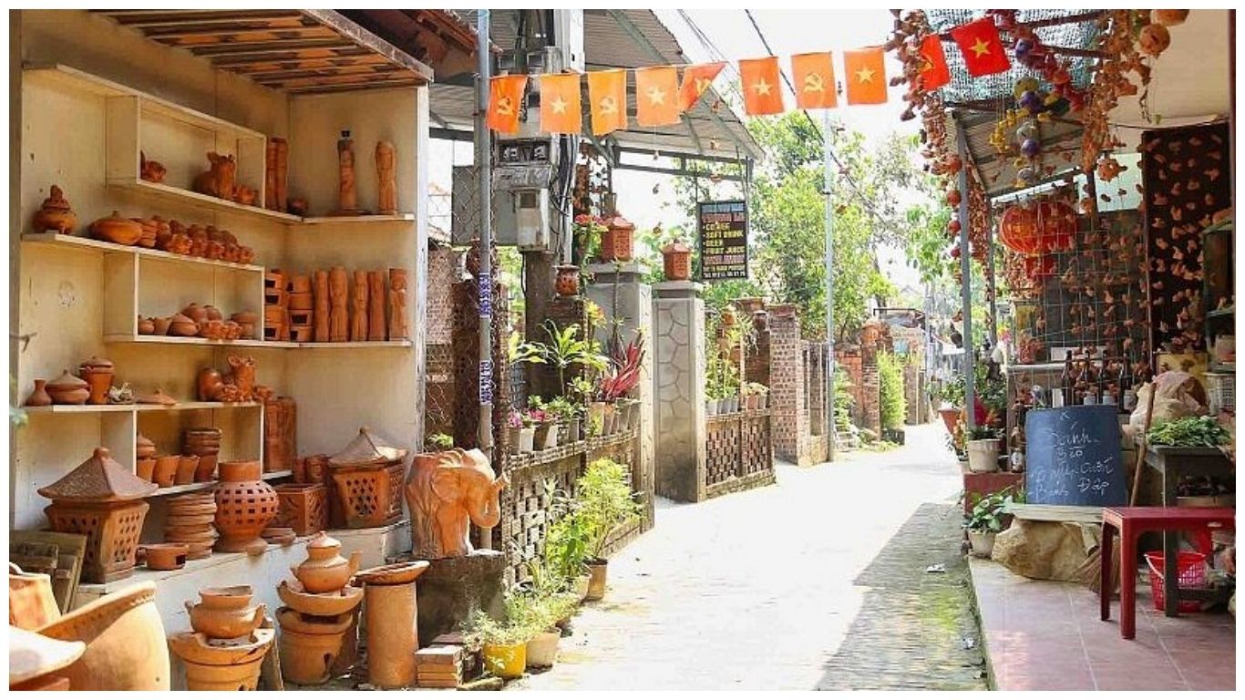 A sunlit alleyway in Thanh Ha Pottery Village Hoi An featuring shelves of traditional terracotta pottery, clay sculptures, and orange Vietnamese flags overhead