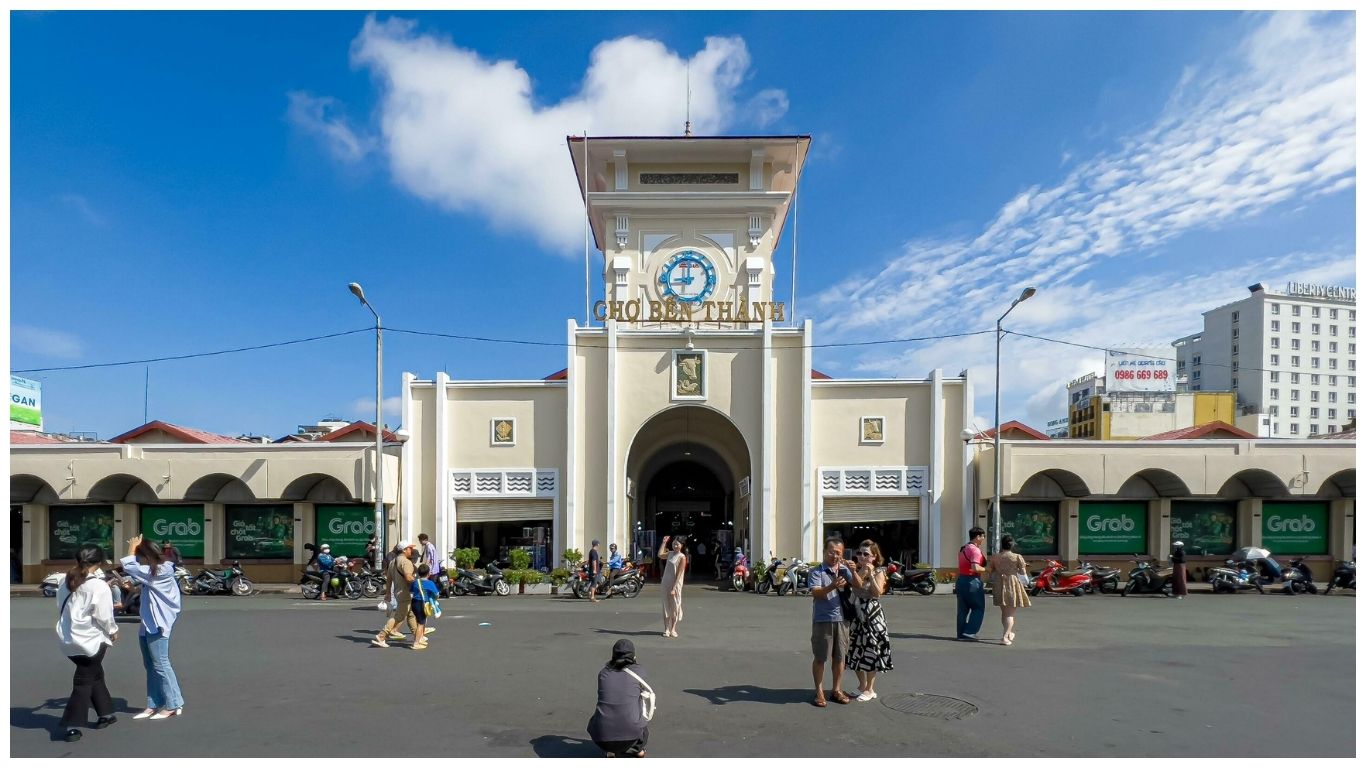 The iconic yellow and white main entrance clock tower of Ben Thanh Market in Ho Chi Minh City under a bright blue sky with tourists and locals in the foreground