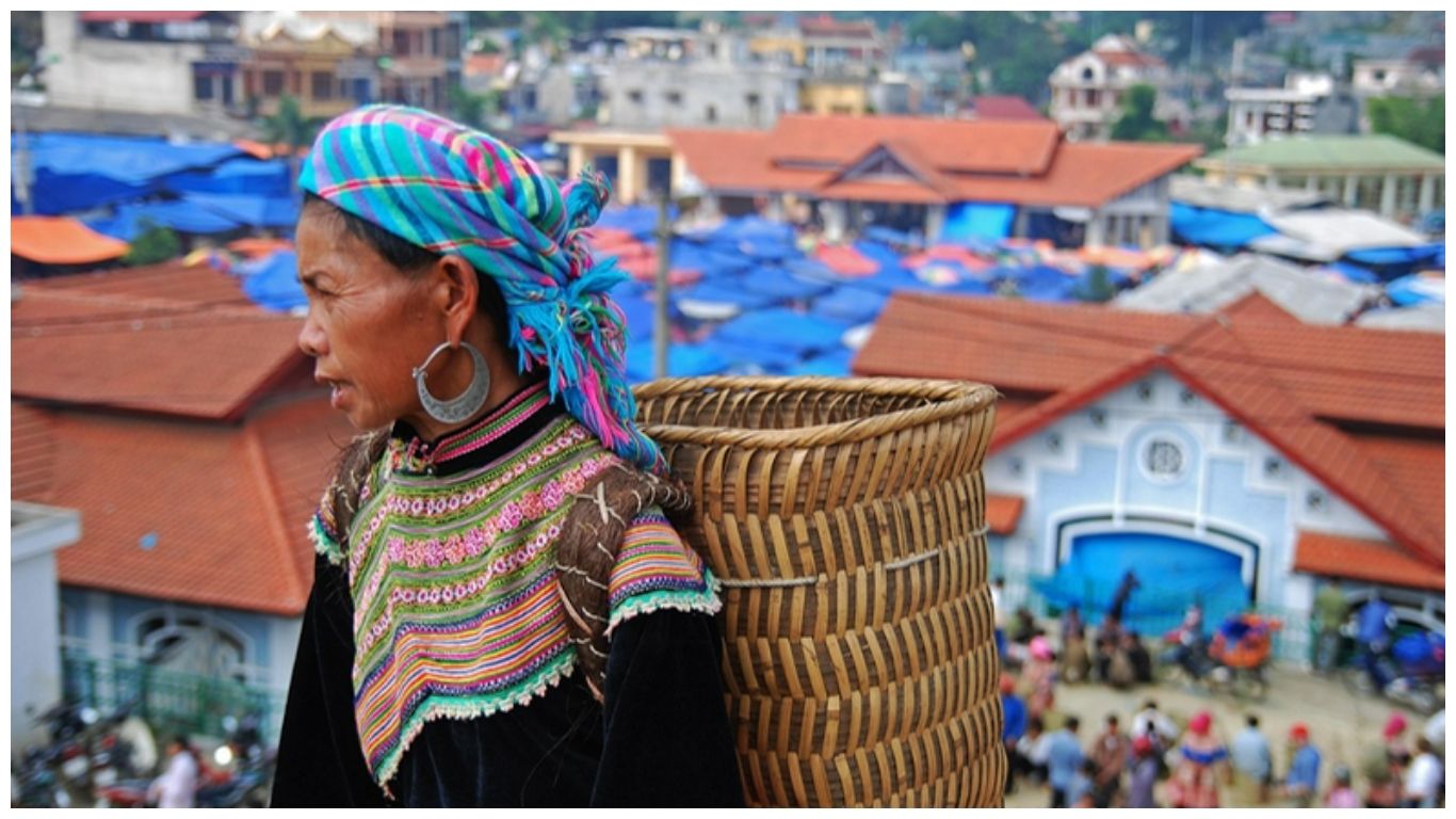 A local Hmong woman in traditional vibrant attire carrying a woven basket overlooking a bustling Sapa market in Northern Vietnam