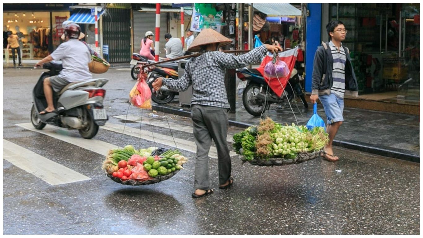 A local street vendor carrying a traditional bamboo pole with baskets of fresh vegetables across a rainy street in Hanoi, Vietnam