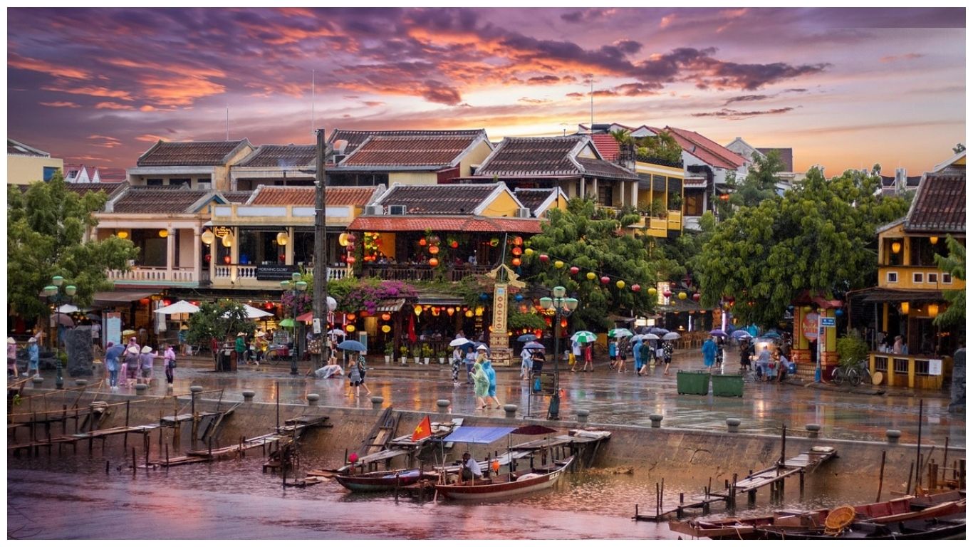 A scenic sunset view of Hoi An Ancient Town in Vietnam featuring traditional yellow buildings, colorful lanterns, and wooden boats on the Thu Bon River during a light rain