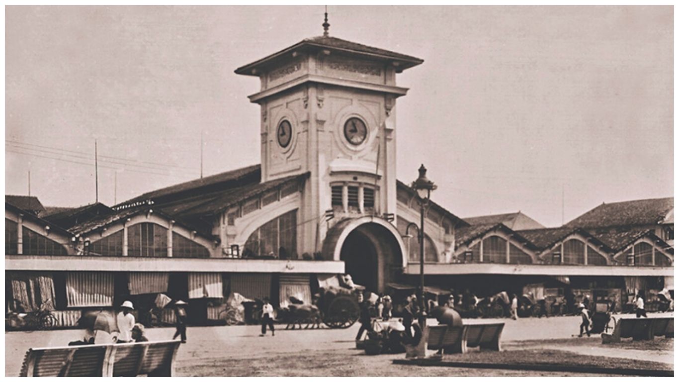 A vintage black-and-white photograph of the historical Ben Thanh Market in Ho Chi Minh City, showing the original clock tower and horse-drawn carriages in the square during the early 20th century