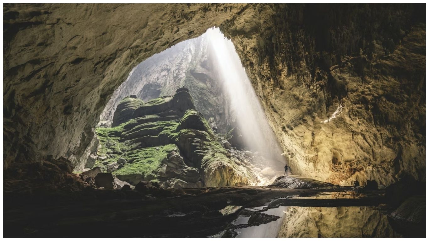 A massive limestone chamber inside Son Doong Cave, Vietnam, featuring a giant doline with sunlight streaming through a roof collapse onto lush green vegetation and a small human figure for scale