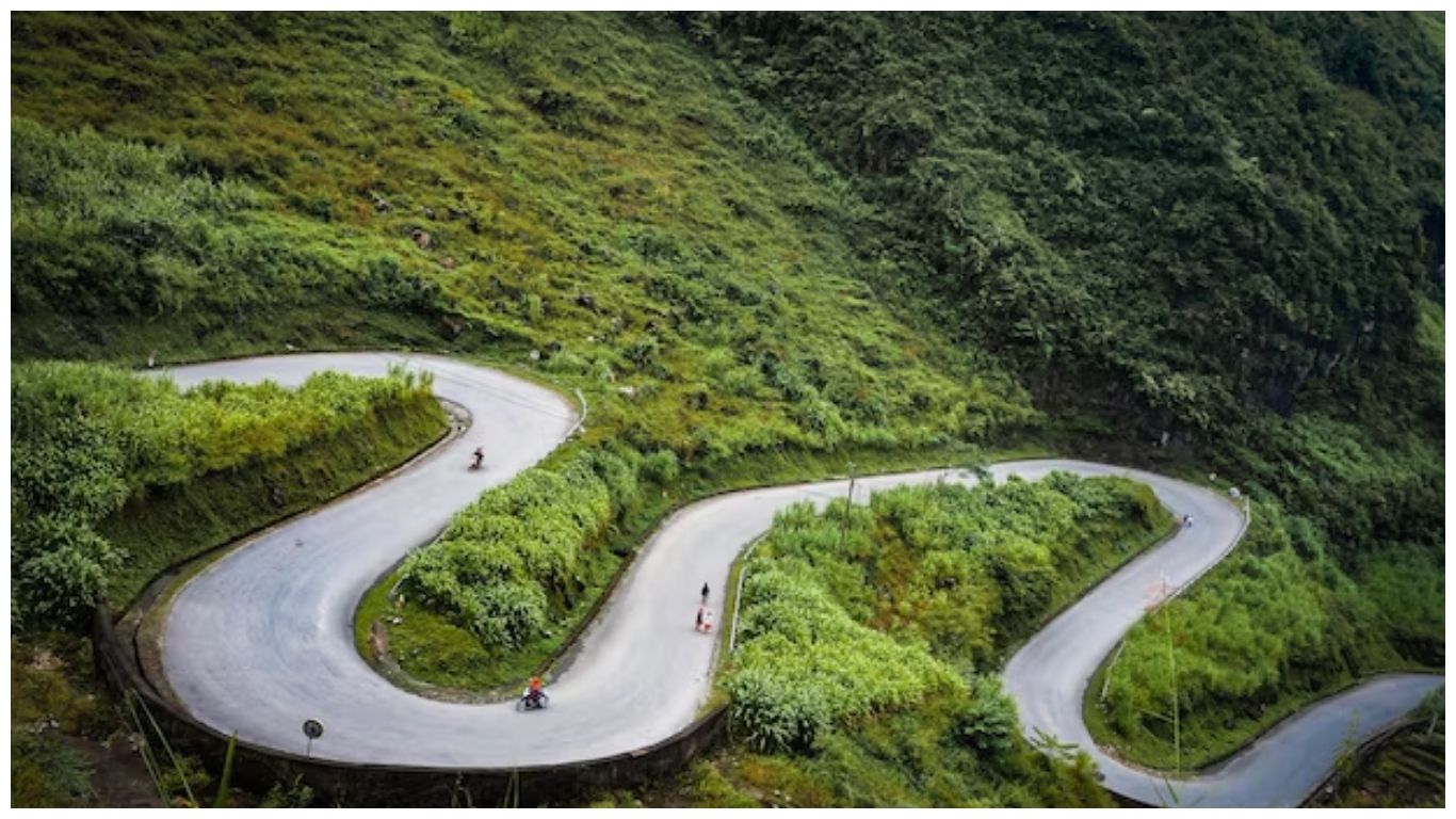 A winding mountain pass on the Ha Giang Loop in Northern Vietnam with motorcyclists navigating sharp curves surrounded by lush green hills
