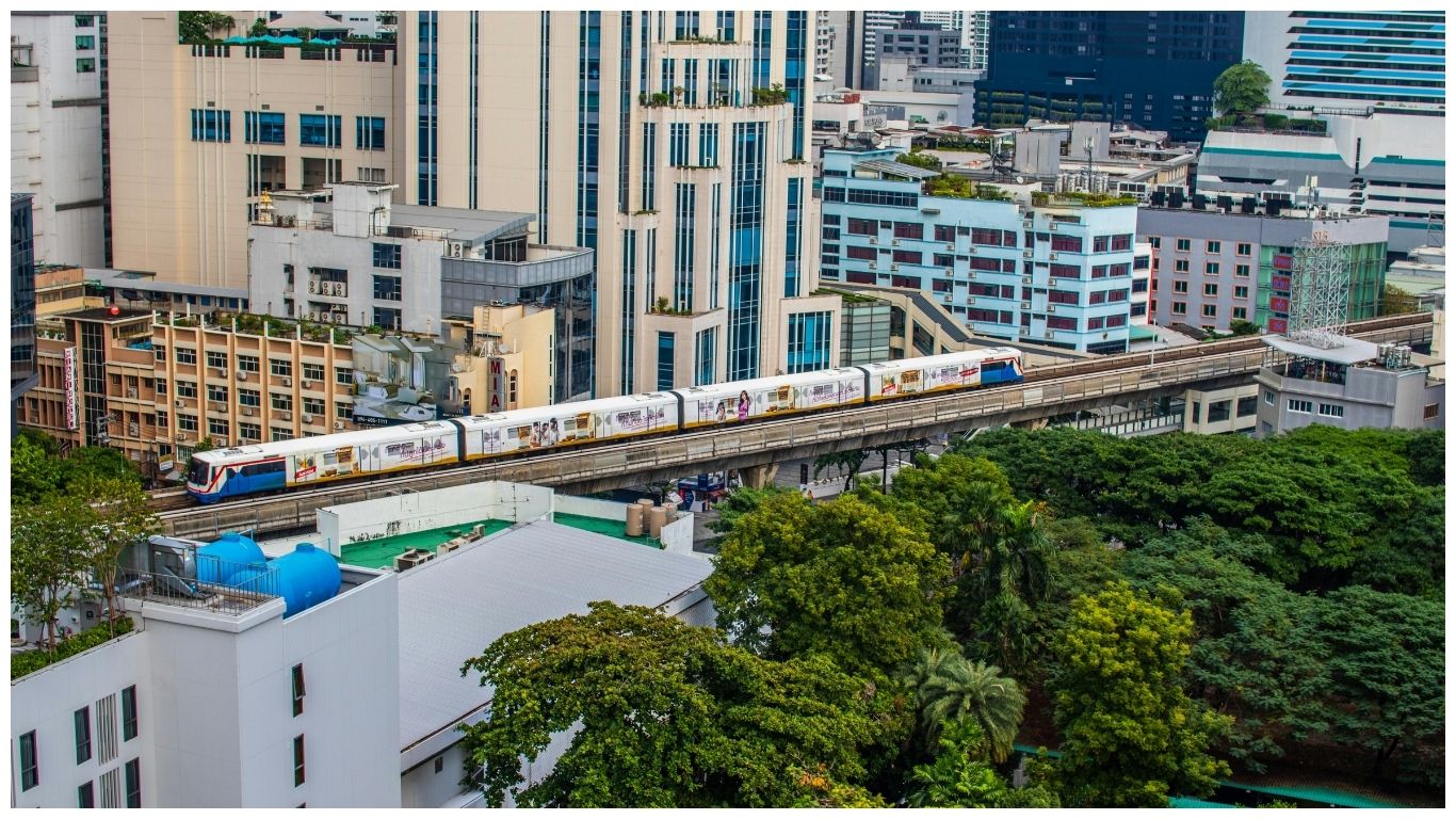 An elevated view of a Bangkok Skytrain (BTS) traveling through the modern urban landscape of Bangkok, surrounded by high-rise buildings and green city trees