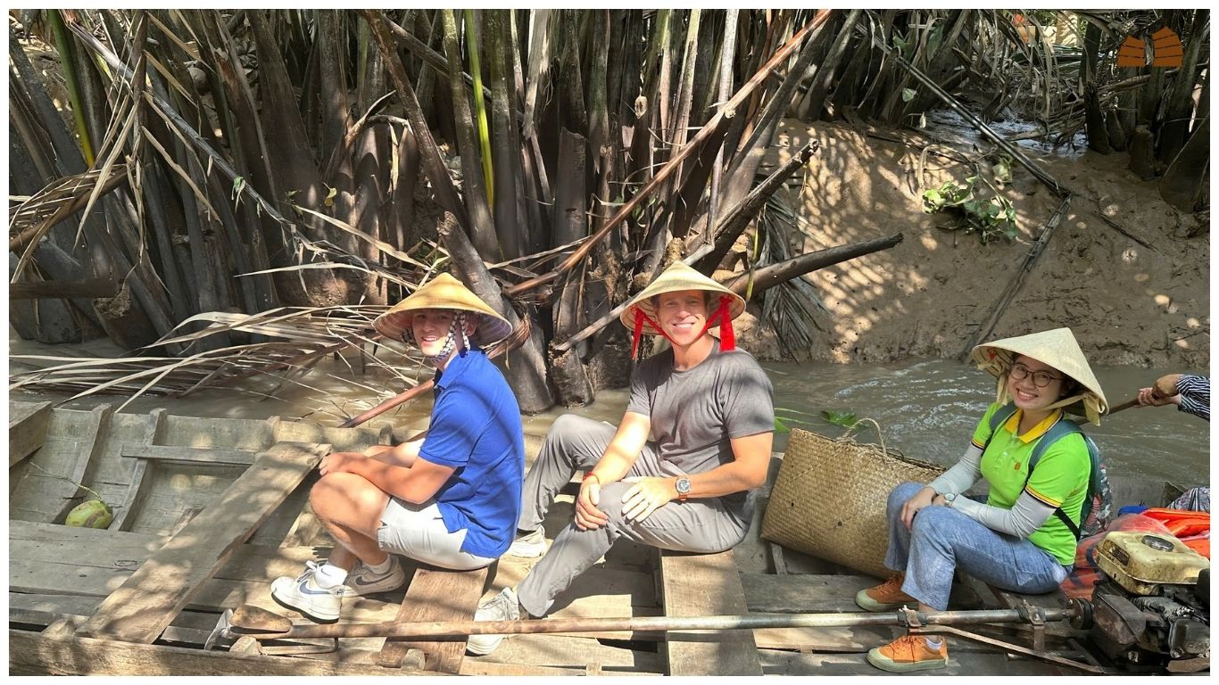Tourists wearing traditional conical hats (non la) on a wooden boat navigating a narrow canal in the Mekong Delta, a top-rated choice for day trips from Saigon