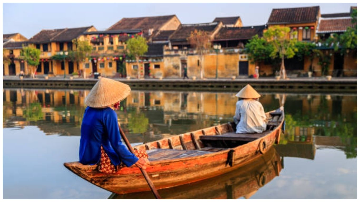 Two local women in conical hats rowing a wooden boat on the Hoai River with ancient yellow houses in the background, offering a peaceful look at the best things to do in Hoi An