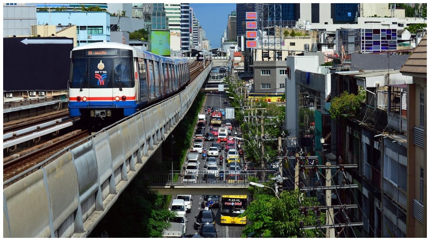 A blue and white BTS Bangkok Skytrain moving on an elevated track directly above a congested city street filled with long lines of cars, buses, and motorcycles in Thailand