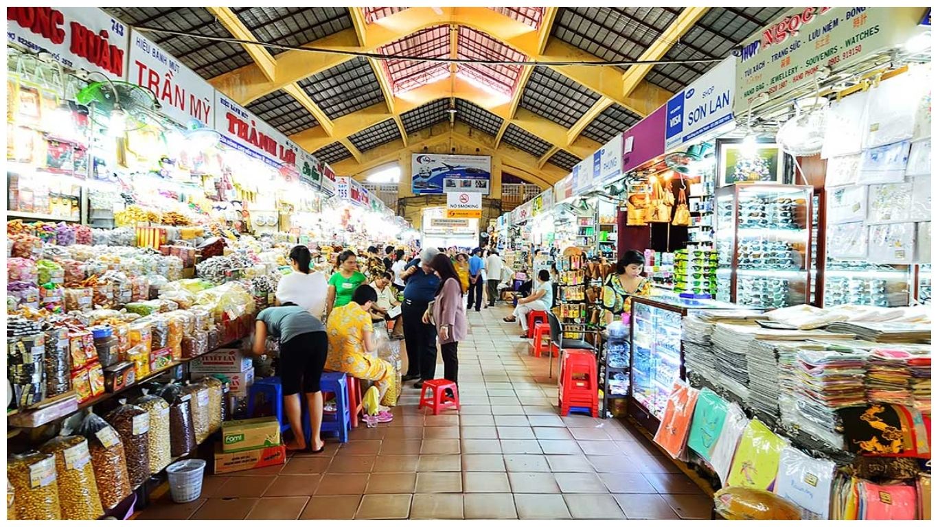 A busy indoor aisle of Ben Thanh Market in Ho Chi Minh City lined with stalls selling dried fruits, nuts, coffee, and colorful souvenirs
