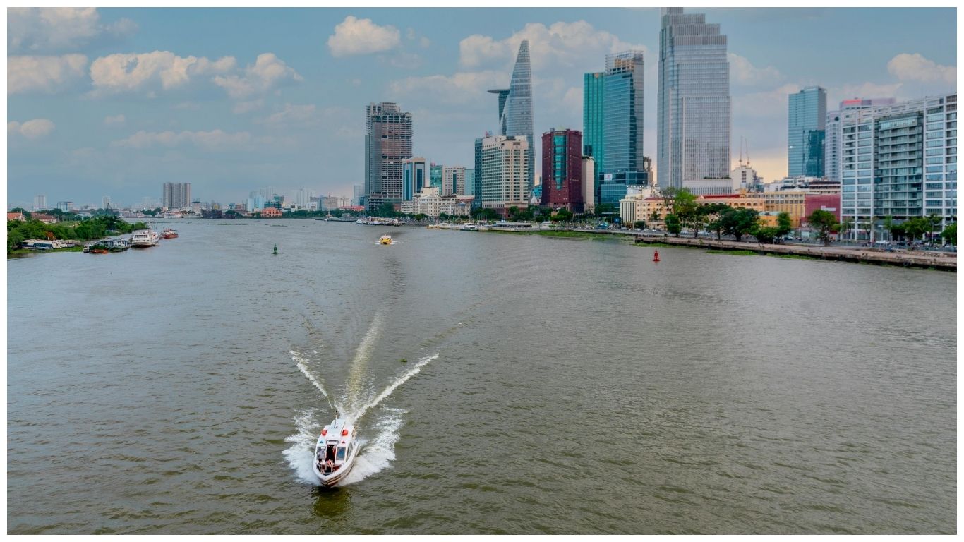 A white speedboat cruising on the Saigon River with the Ho Chi Minh City skyline and Bitexco Tower in the background, a scenic start to day trips from Saigon