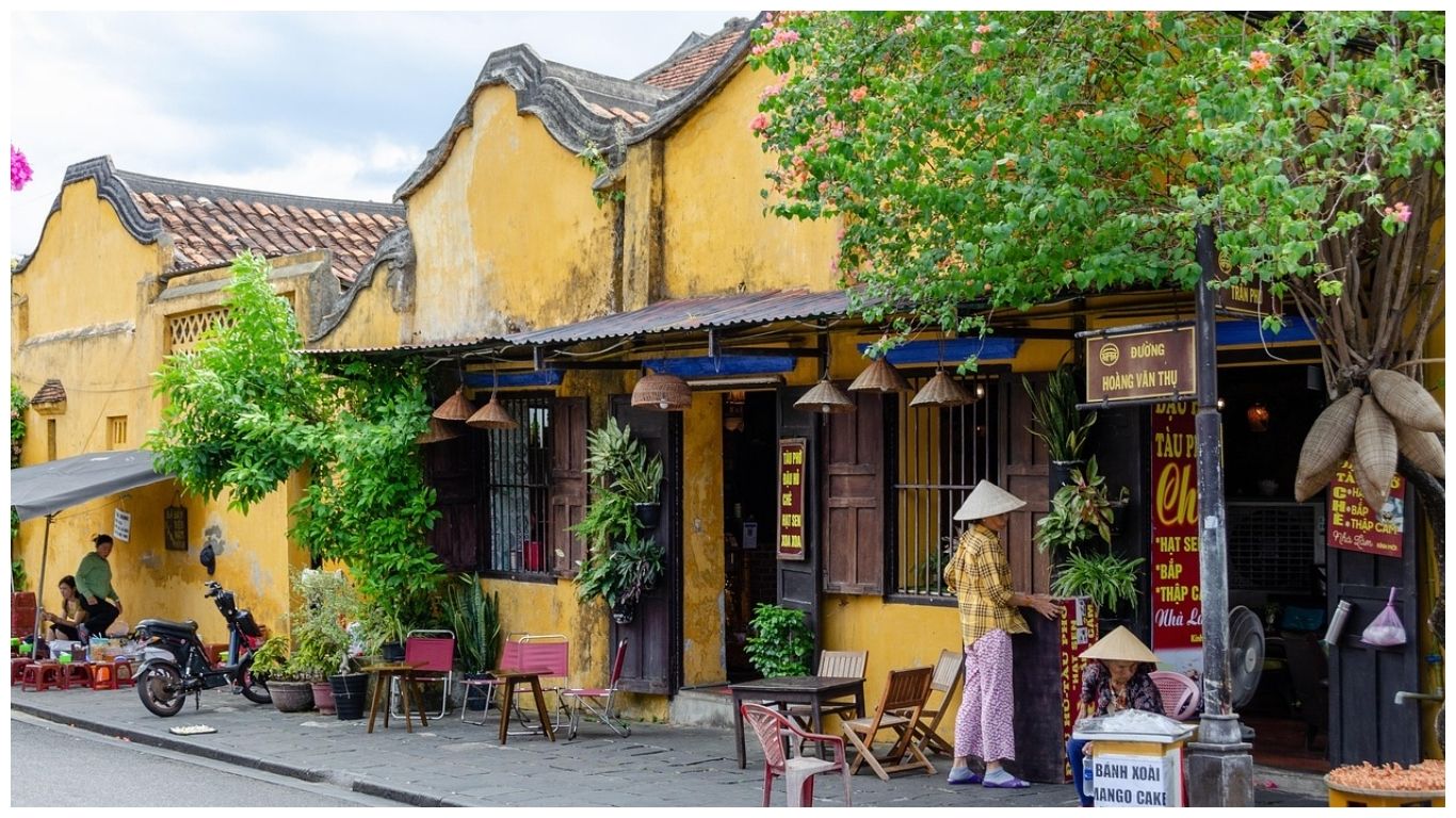 A local street scene in Hoi An Ancient Town featuring a yellow heritage building, a small sidewalk café, and locals in traditional conical hats