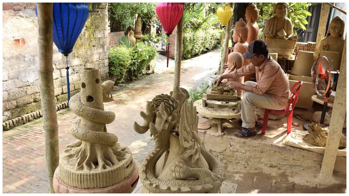A skilled local artisan at Thanh Ha Pottery Village Hoi An carefully hand-carving intricate details into a large clay dragon sculpture under the shade of colorful traditional lanterns
