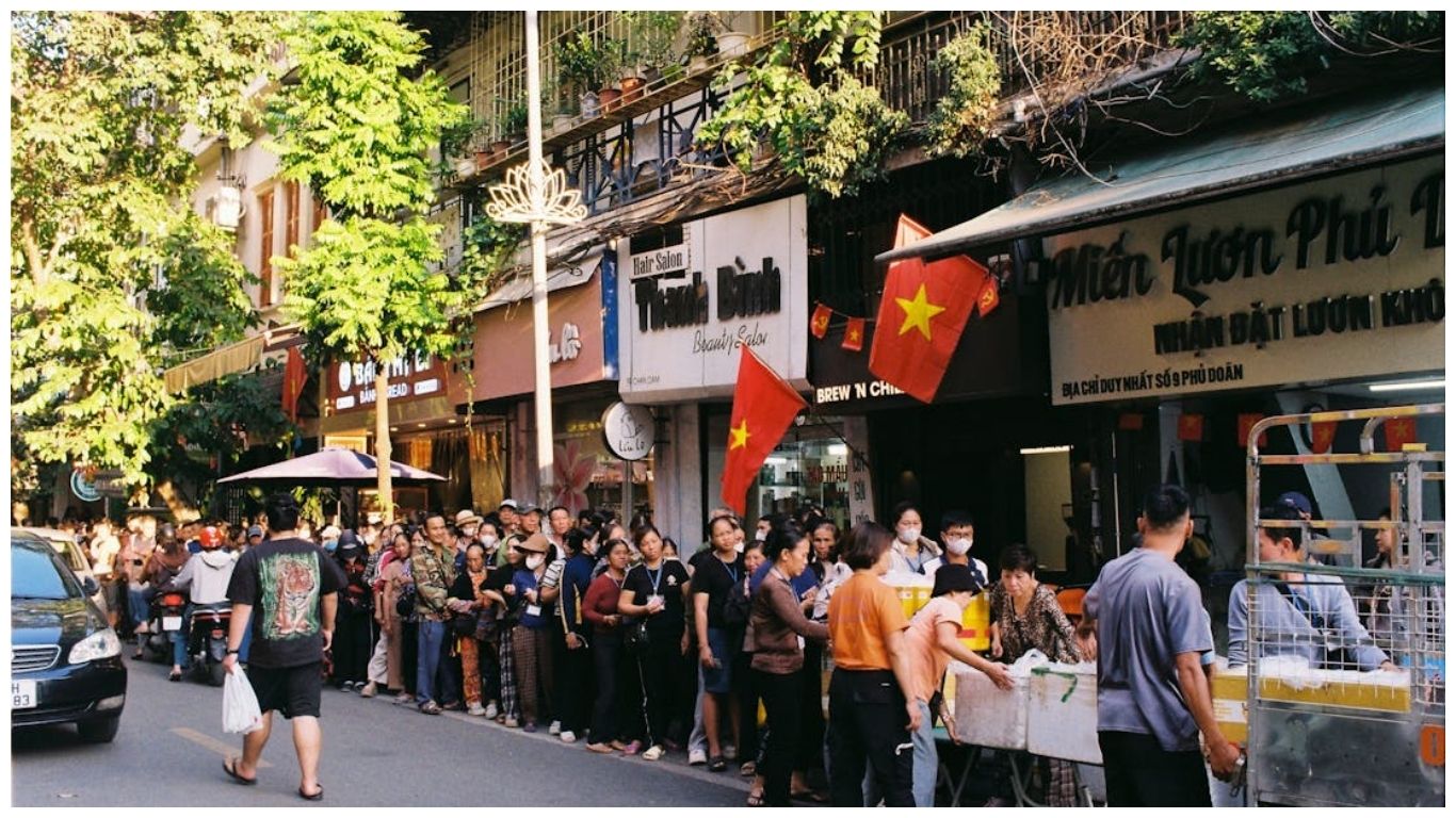 A long queue of locals and tourists on a busy Hanoi street with Vietnamese flags, showcasing the vibrant local life and street food culture