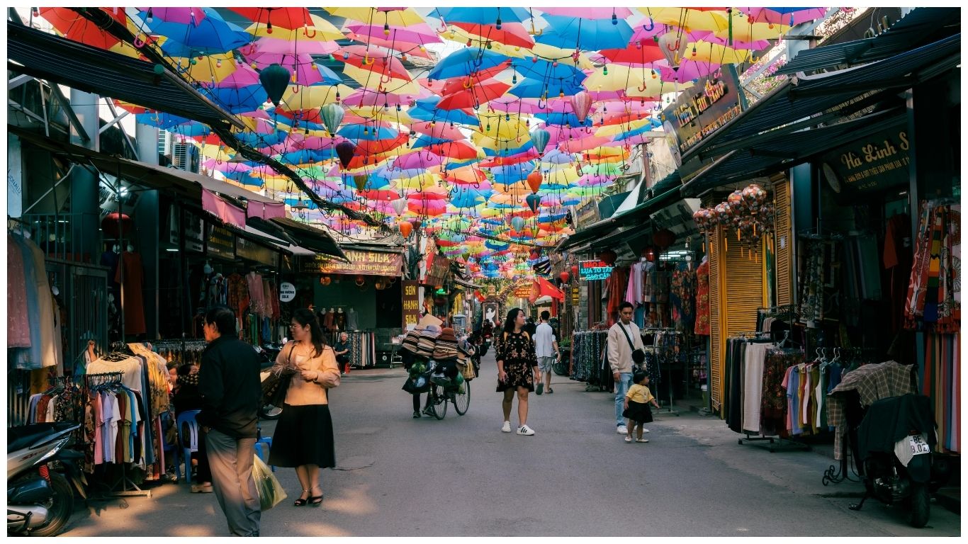 A bustling narrow shopping street in Vietnam decorated with hundreds of colorful umbrellas and lanterns hanging overhead, with local shoppers and vendors walking past clothing stalls