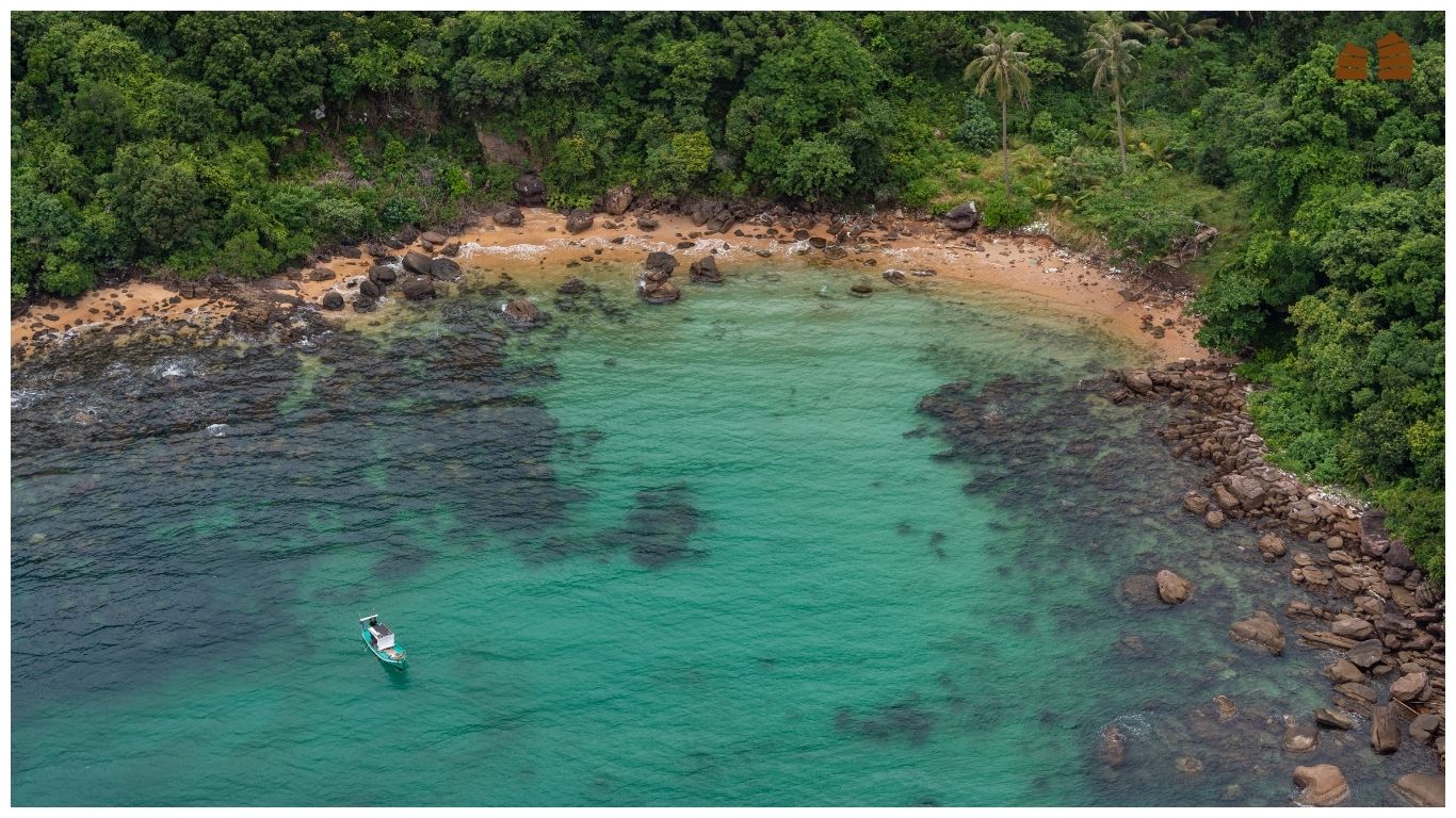 An aerial drone view of a small turquoise bay in Phu Quoc with lush green jungle, rocky shores, and a single traditional fishing boat anchored in the clear water