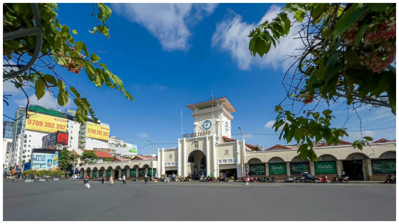 A wide view of the cream-colored Ben Thanh Market in Ho Chi Minh City clock tower under a blue sky, framed by green leaves with the modern city skyline and billboards in the background