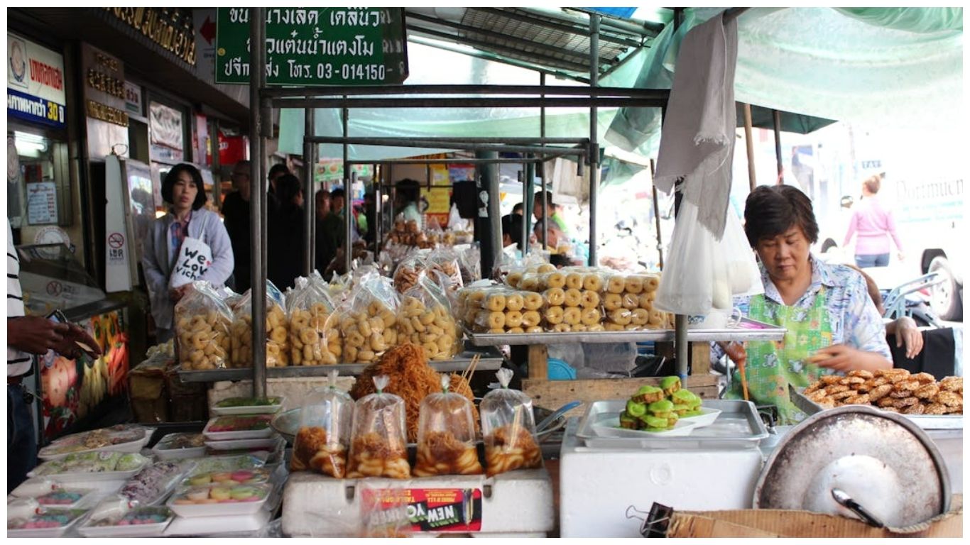 A busy local street food stall in Bangkok featuring a variety of Thai snacks and desserts, perfect for travelers deciding between Hanoi or Bangkok