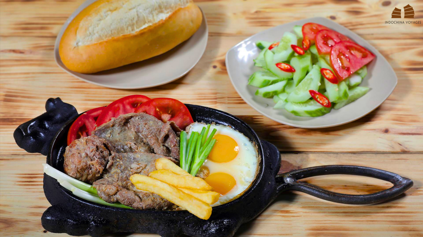 Hot plate of bo bit tet with beef, two fried eggs and fries, served with bread and cucumber salad in Hanoi